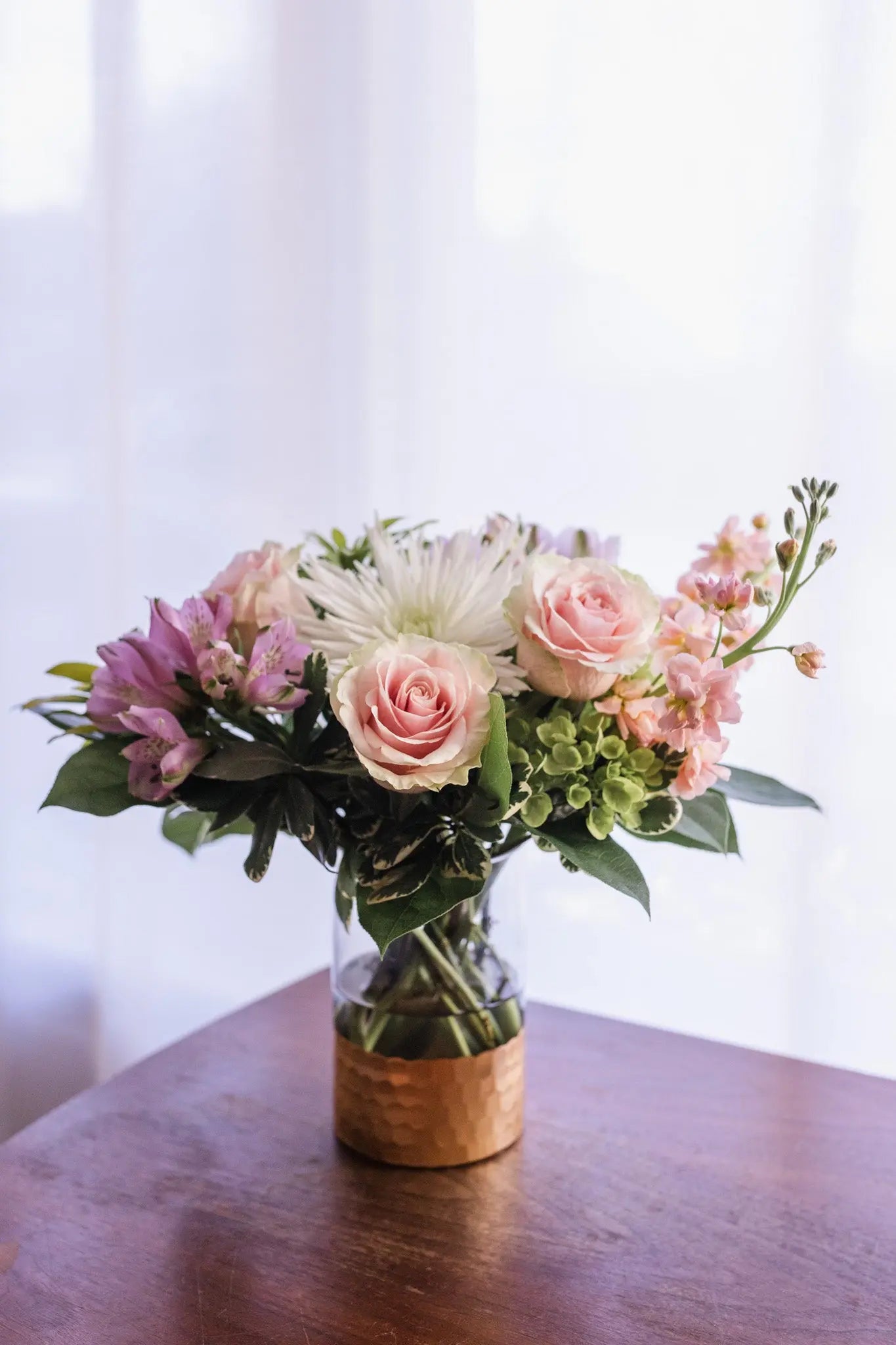A floral arrangement featuring pastel colors, with roses and other mixed flowers, in a clear glass vase, placed on a wooden surface.