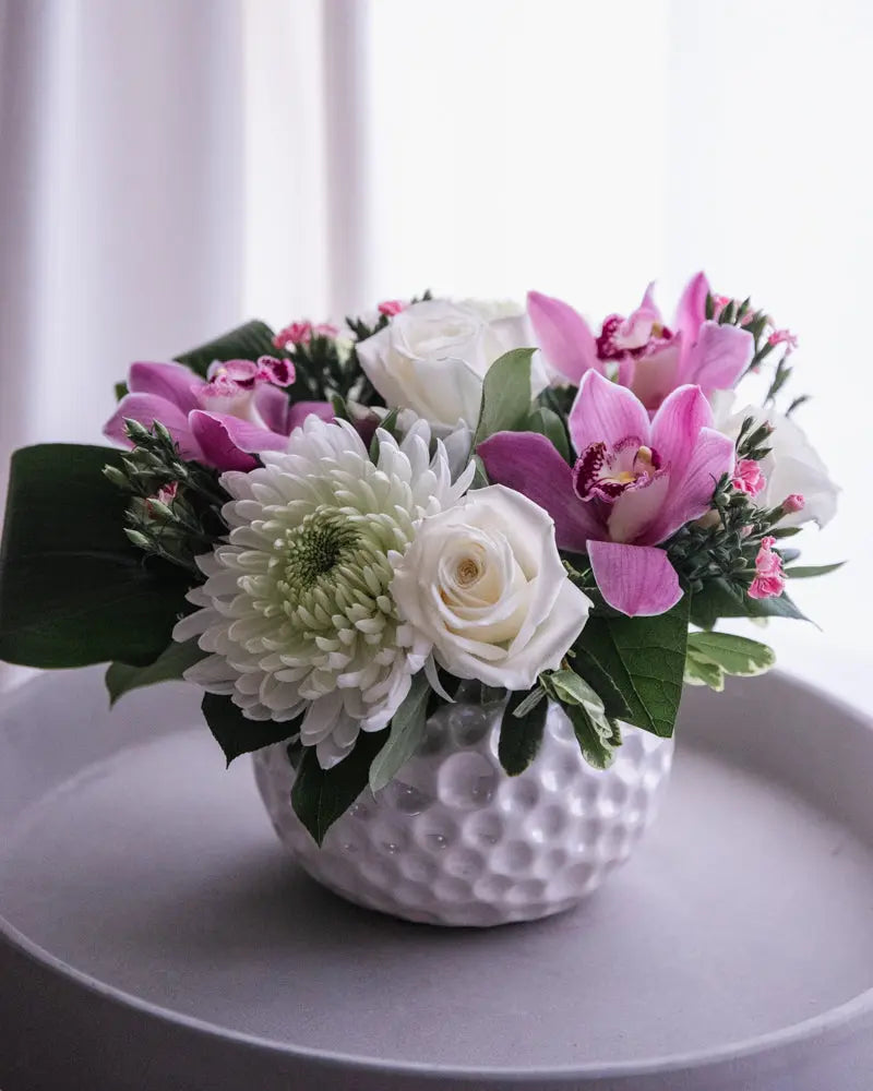 A floral arrangement featuring white roses, Cymbidium orchids, and various greenery in a white textured vase.