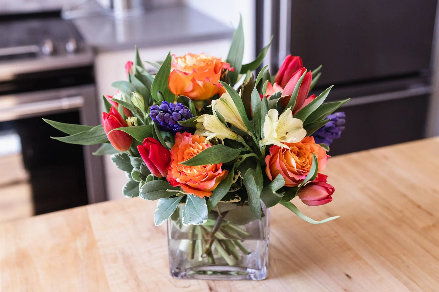 A glass cube vase containing a colorful floral arrangement of Hyacinths, Tulips, Roses, and Alstromeria.