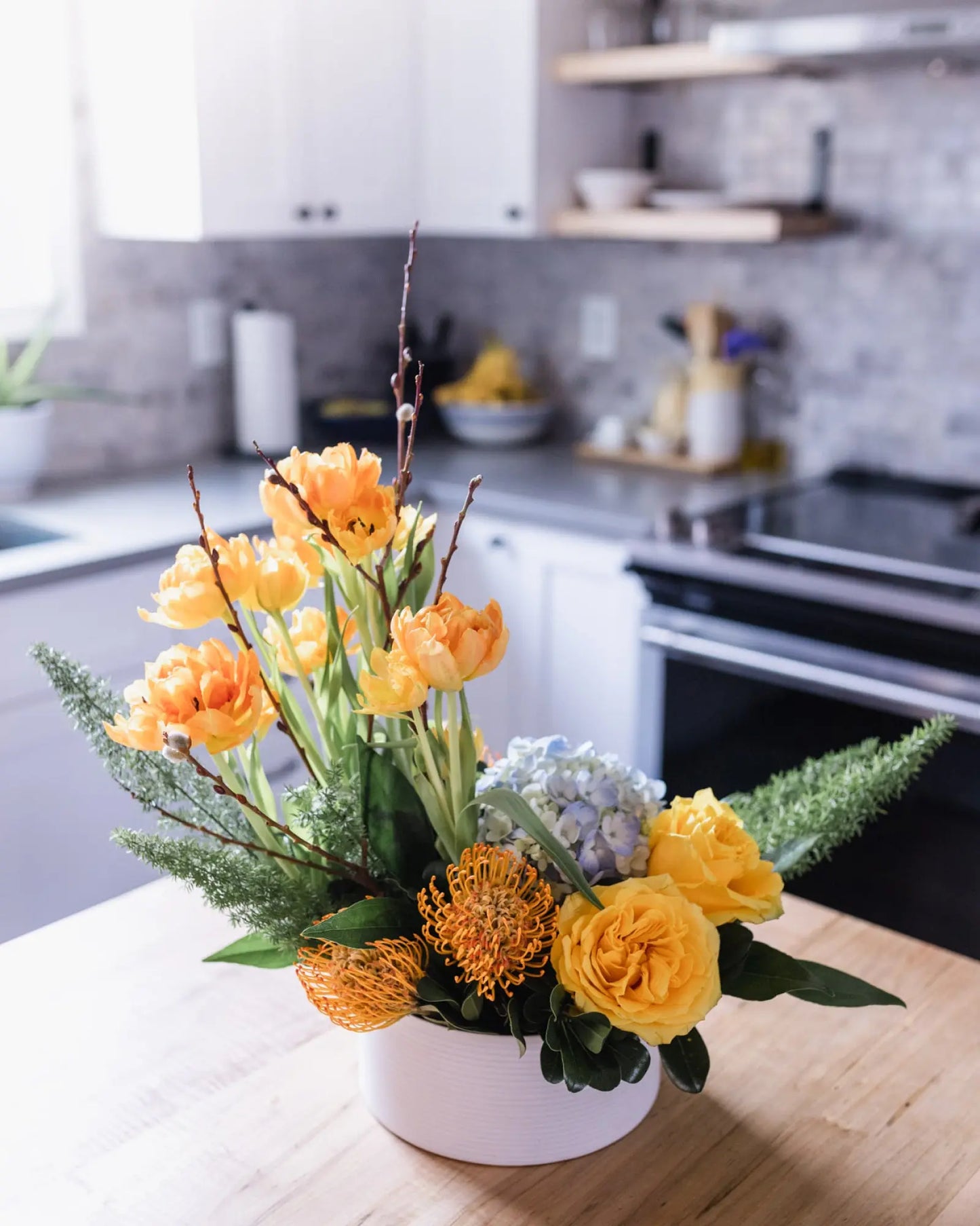 A floral arrangement featuring double tulips, pussy willow, yellow roses, and blue hydrangea in a white container.