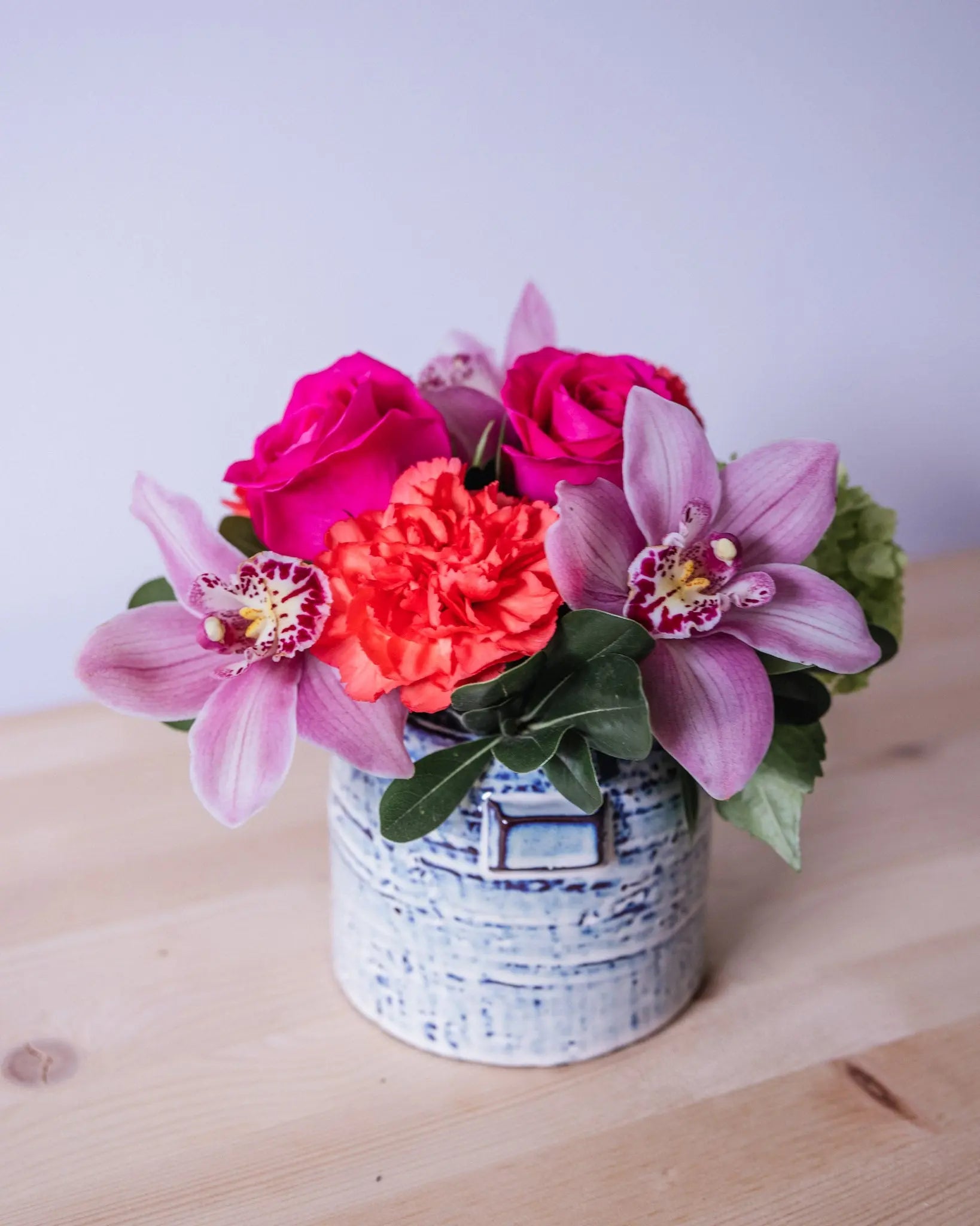 A floral arrangement featuring hot pink roses, cymbidium orchids, and orange carnations in a ceramic vase.