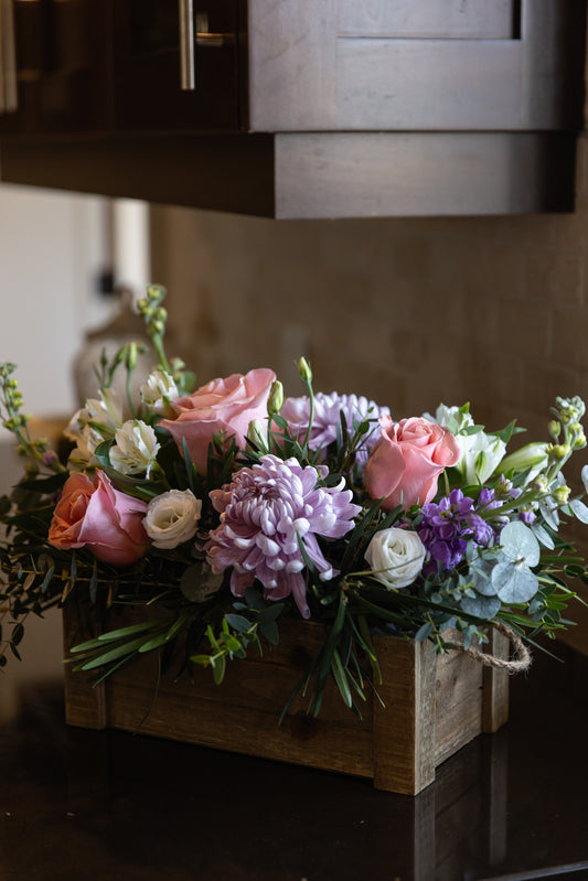 A floral arrangement featuring a variety of flowers including roses and stock in colors pink, purple, white, and green, displayed in a wooden box.