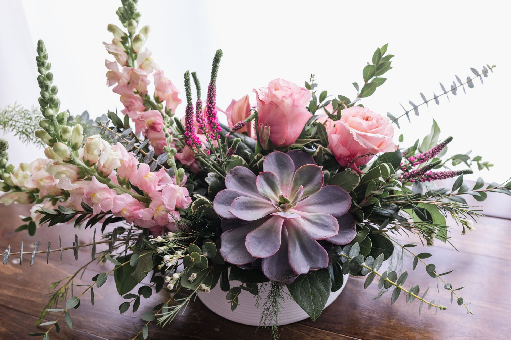 A floral arrangement featuring soft pink blooms, snap dragons, roses, orchids, and a succulent in a white container, placed on a wooden surface.