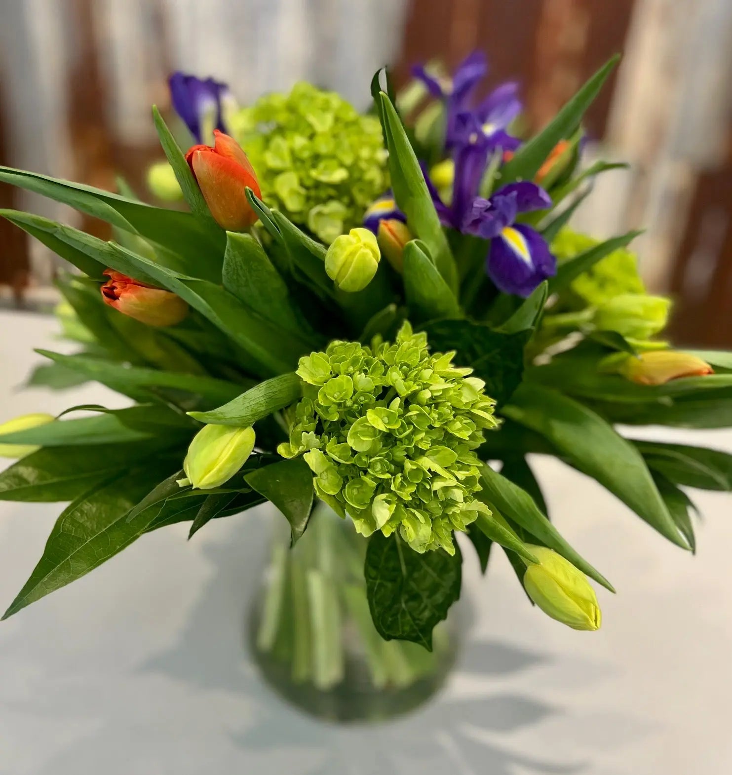 A floral arrangement featuring a mix of tulips, purple irises, and green hydrangea in a clear glass vase,
