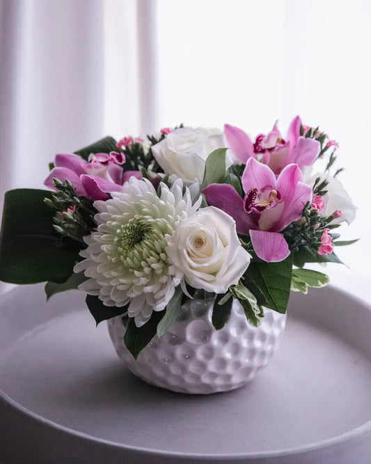 A floral arrangement featuring white roses, Cymbidium orchids, and various greenery in a white textured vase.